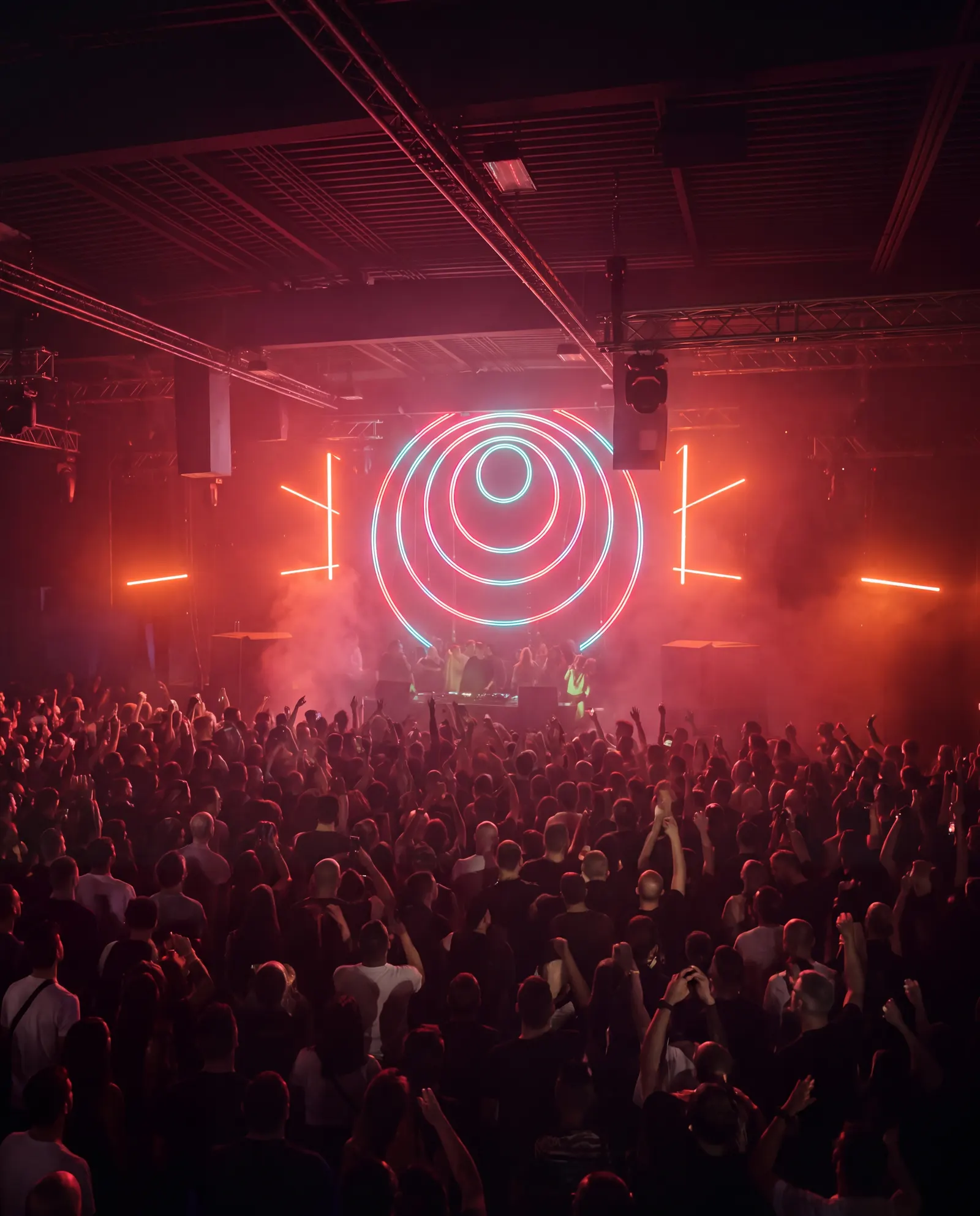 A crowd with hands raised under concentric neon rings at Demo Room, red haze across the stage
