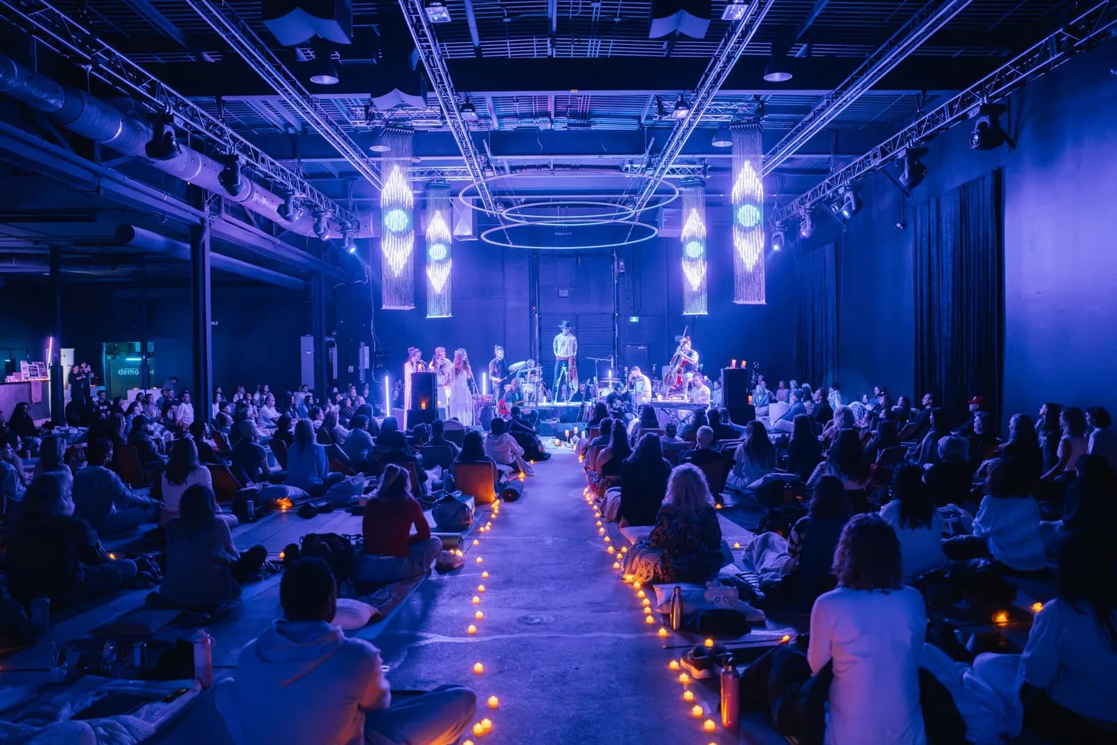 A seated audience on mats facing a lit stage in Demo Room