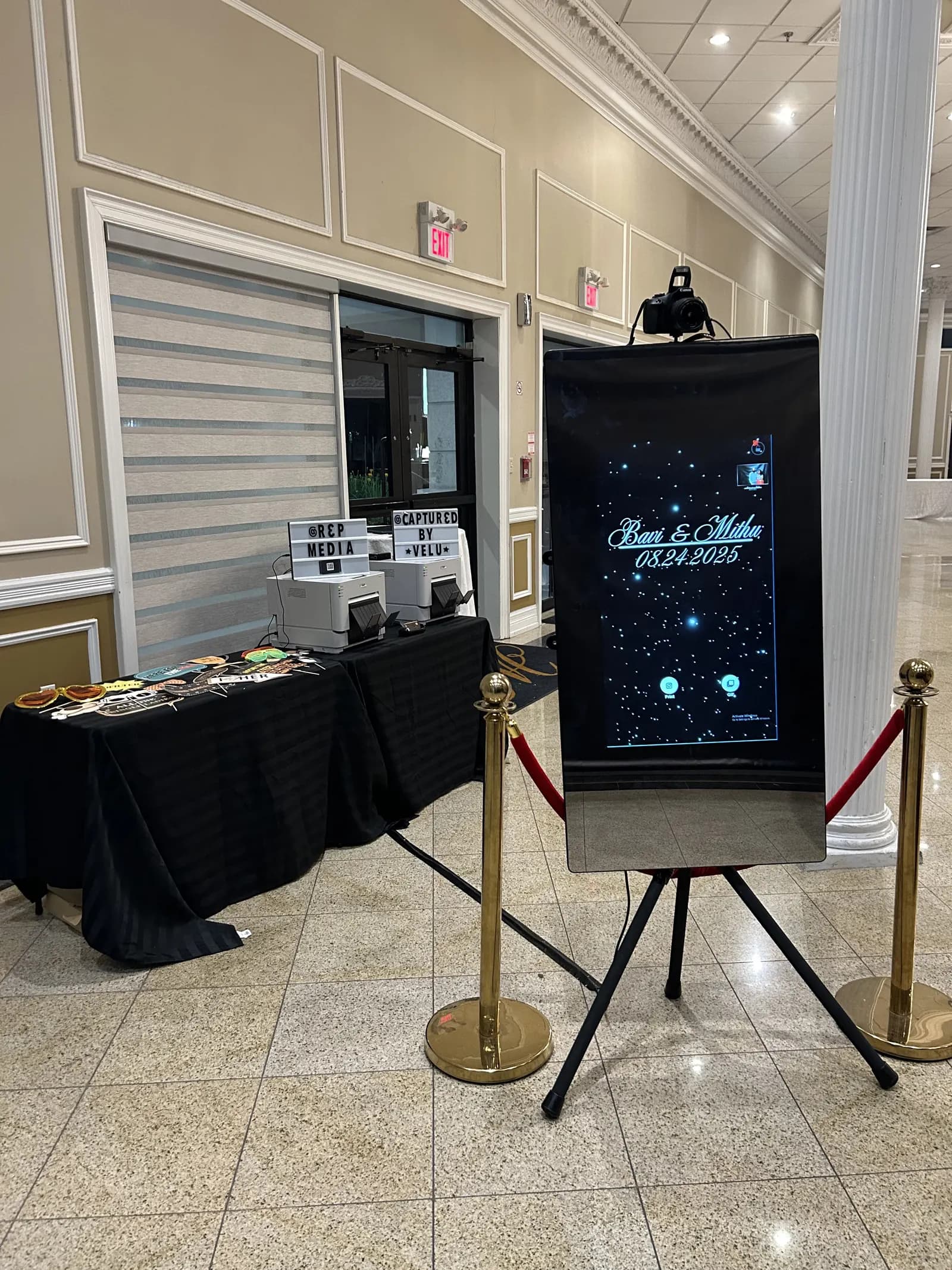 A touch-screen photo booth on an easel with a print station, props table and rope line
